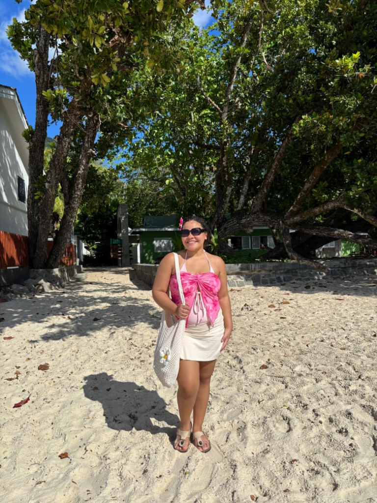 A young woman walking on a sunny beach wearing a pink top and white skirt, surrounded by trees and soft sand.