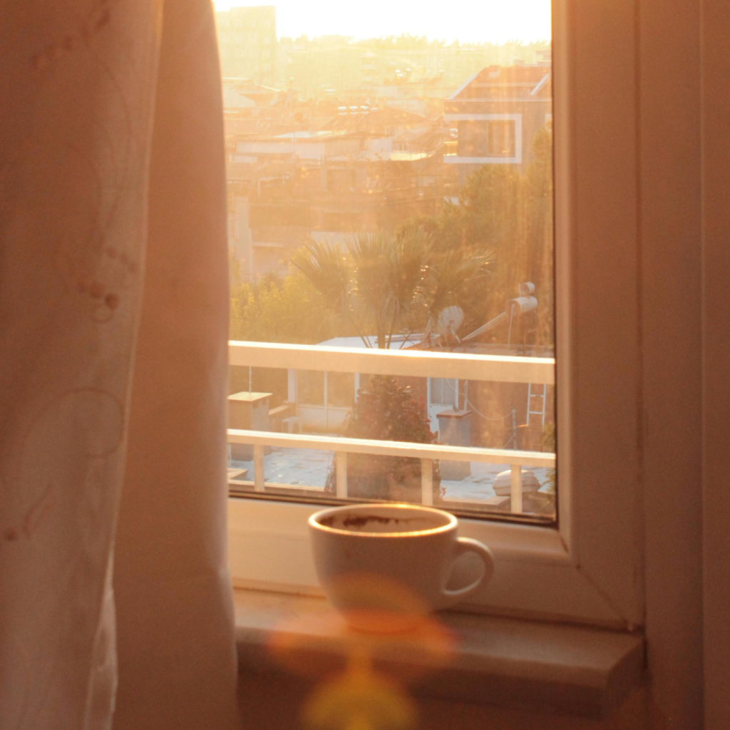A warm mug resting on a windowsill as soft golden sunlight filters through sheer curtains at sunset.