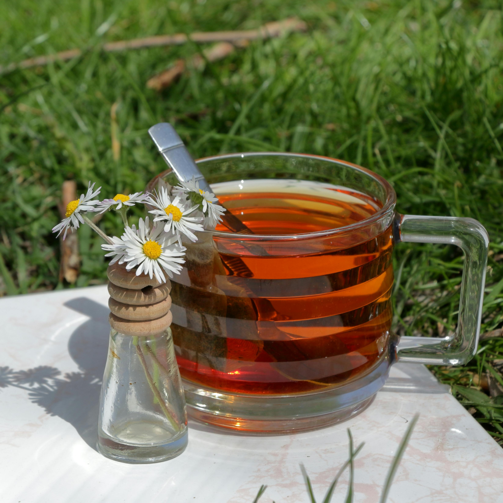 A clear glass mug of warm herbal tea with a silver spoon beside a small bottle of white wildflowers, placed outdoors on grass in soft natural light.